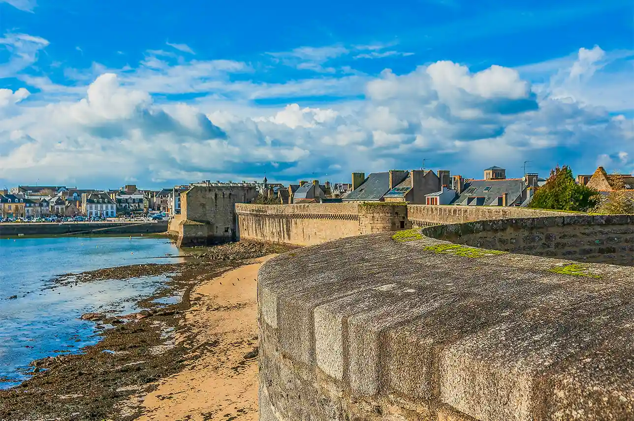 Vue panoramique des remparts de la Ville Close de Concarneau, surplombant la plage et le port par une journée ensoleillée avec ciel bleu et nuages bretons.