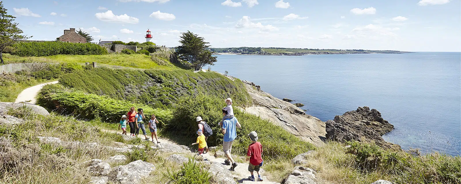 Famille en randonnée sur le sentier du GR34 à Concarneau, longeant la côte bretonne avec vue sur la mer et un phare rouge au loin.